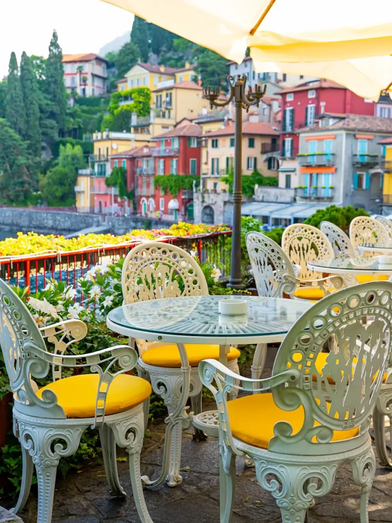 Ornate white metal café tables and chairs with yellow cushions on a terrace overlooking Lake Como Italy with colorful hillside buildings and lush greenery in the background.