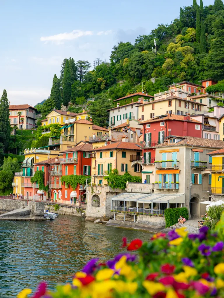 The Perfect Milan To lake Como Day Trip (Self-Guided) colourful lakeside buildings along Varenna A peaceful waterfront scene on Lake Como Italy with warm toned buildings set against a lush green mountainside.