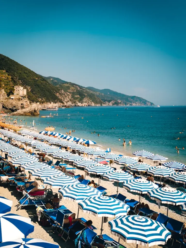Panoramic view of Monterosso Beach in Cinque Terre, Italy, showing turquoise sea, golden sandy shore, colorful rows of blue and yellow umbrellas, and a mountain backdrop under a clear sky