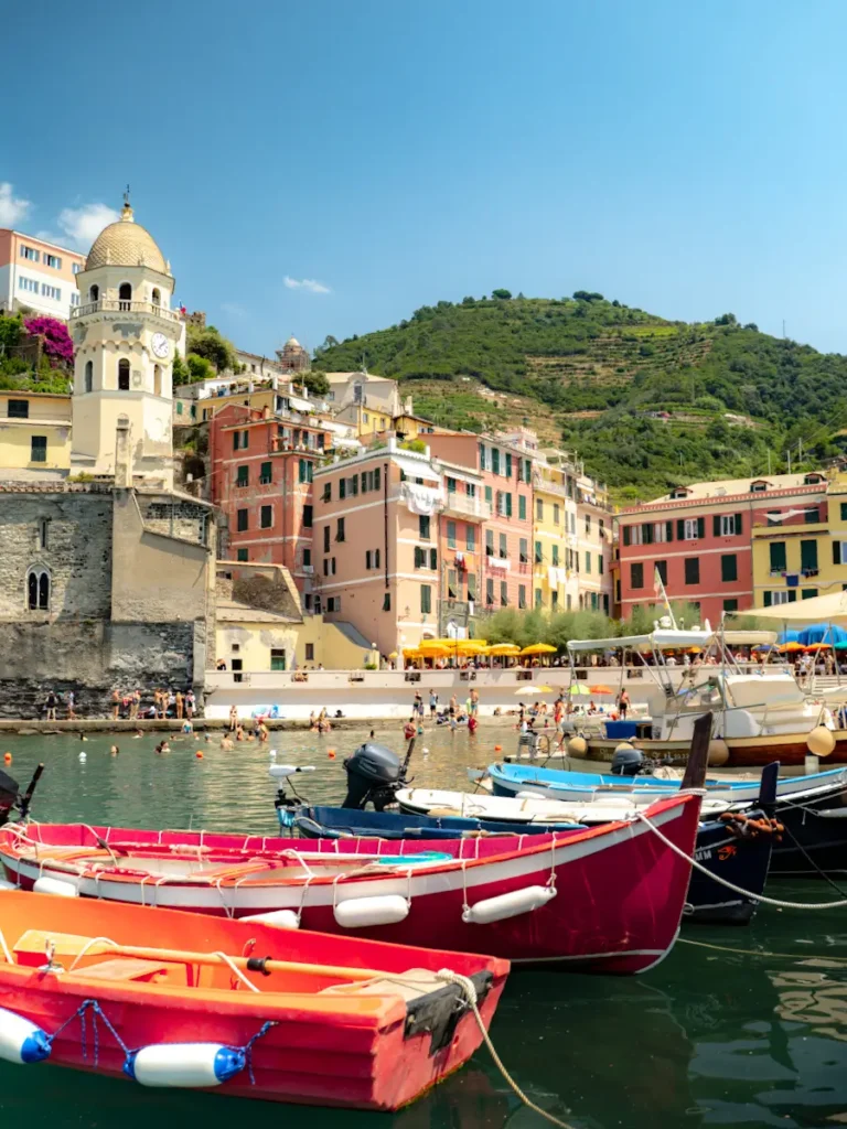 a view of Vernazza harbour with colourful buildings in the background and red boats in the foreground and a view of a green hill in cinque terre italy