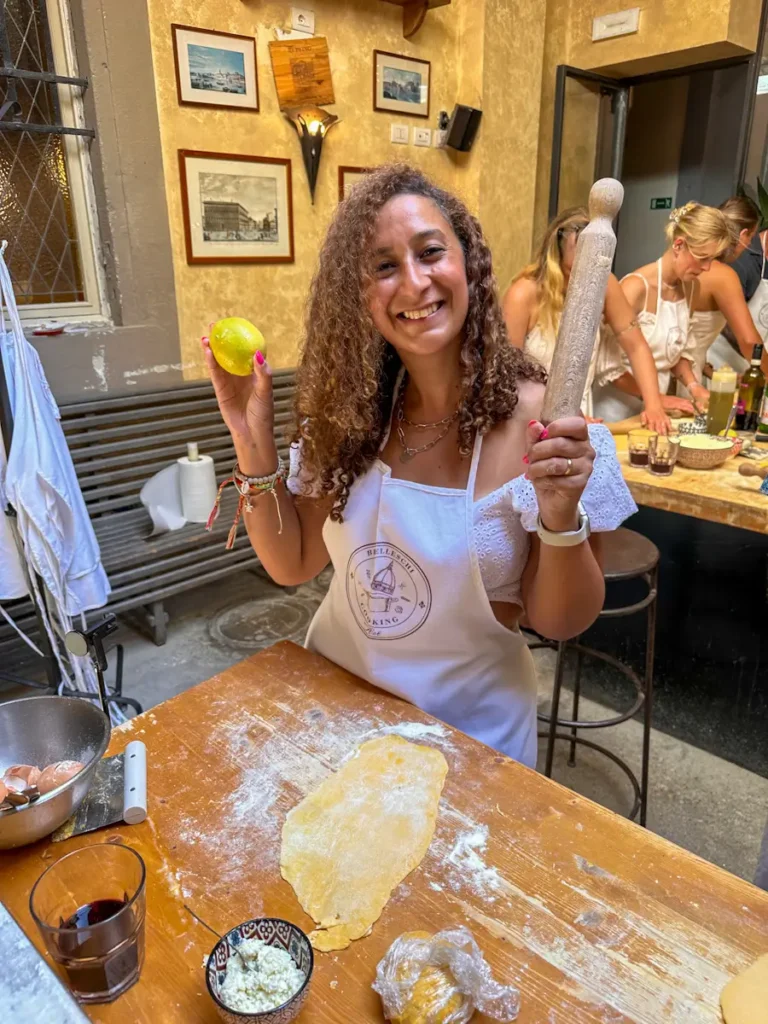 Sally Elia is smiling during a Florence, Italy cooking class, holding fresh pasta dough in her hand with another piece of dough on the table, surrounded by ingredients and a lively classroom