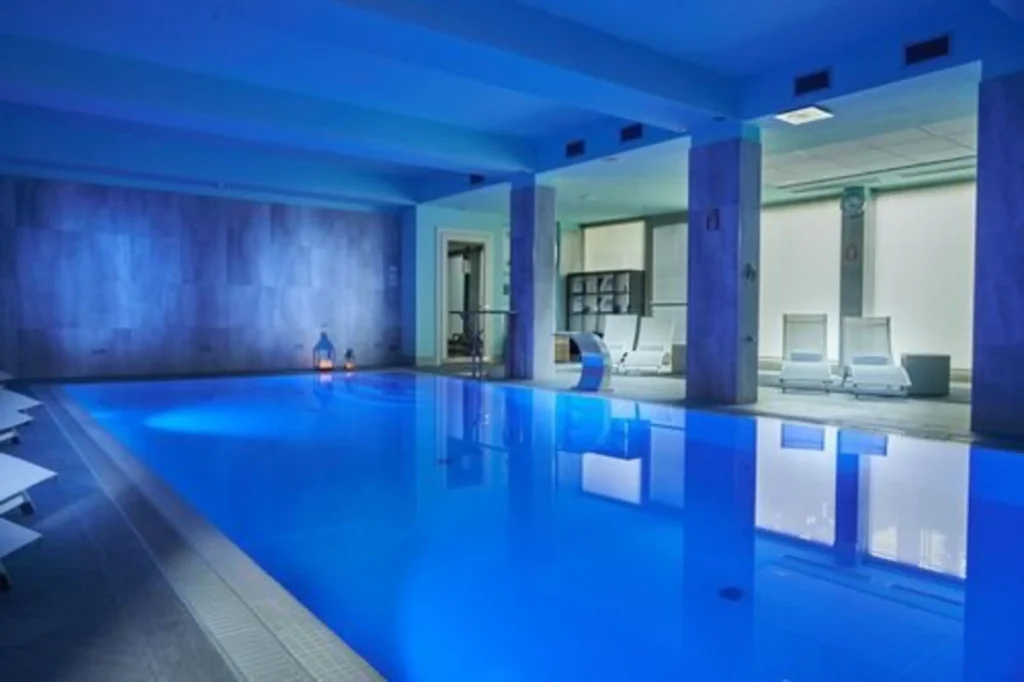 Indoor pool inside Forte 16 hotel in florence italy, the pool is lit with deep blue lighting reflecting off the water and surrounding walls. Glass doors and clean lines create a modern spa like atmosphere.