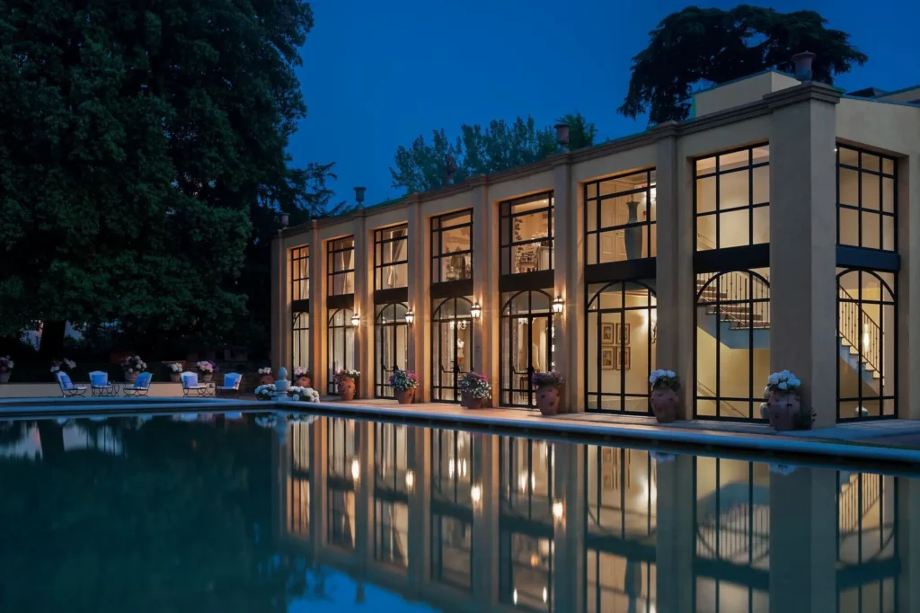 Multi story building in Four Seasons Hotel Firenze with large glass windows glowing warmly at night. Outdoor seating and outdoor luxury swimming pool is visible below the structure adding to the inviting ambiance, Four Seasons Hotel Firenze is one of the hotels in Florence Italy with a pool

