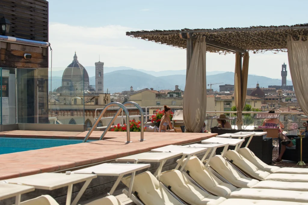 Rooftop terrace in Hotel Kraft Firenze Italy with an outdoor swimming pool overlooking Florence cityscape with the cathedral dome visible above the buildings. White lounge chairs are arranged around the pool