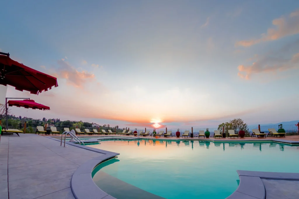 Infinity pool in Villa Tolomei hotel Florence italy overlooking a wide body of water at sunset with soft orange and pink hues in the sky. The pool edge blends seamlessly with the horizon, White lounge chairs are arranged neatly along the terracotta deck