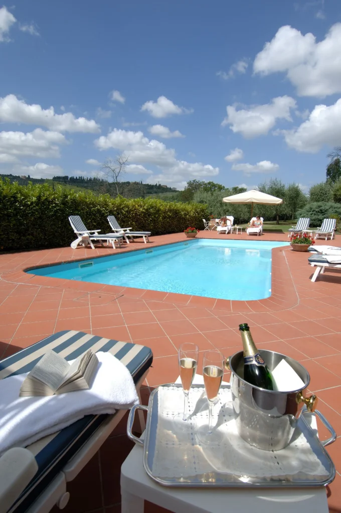 Rooftop terrace in Marignolle Relais & Charme hotel in florence italy with a rectangular swimming pool overlooking rolling green hills under a partly cloudy sky. White lounge chairs are arranged neatly along the terracotta deck