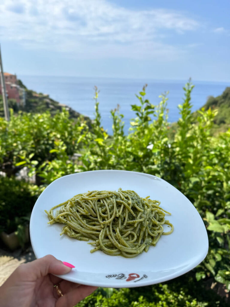 Best Cinque Terre Tour From Florence: Review & My Experience Plate of traditional pesto pasta with lush green hills, blue sea views, and sunny skies in Cinque Terre Italy