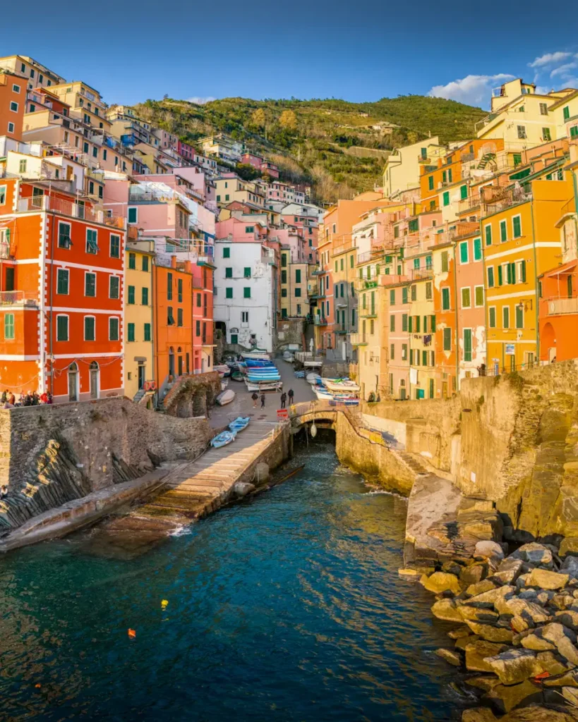 Best Cinque Terre Tour From Florence: Review & My Experience Narrow street lined with warm-toned pastel buildings, outdoor cafรฉs, and bright summer skies in Riomaggiore, Cinque Terre Italy