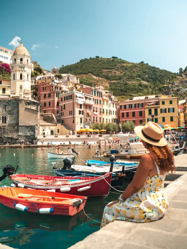 Best Cinque Terre Tour From Florence: Review & My Experience Woman sitting by the turquoise water and Red fishing boats floating beside pastel buildings and crystal-clear water in Vernazza harbour, with colourful cliffside buildings and a bright blue sky